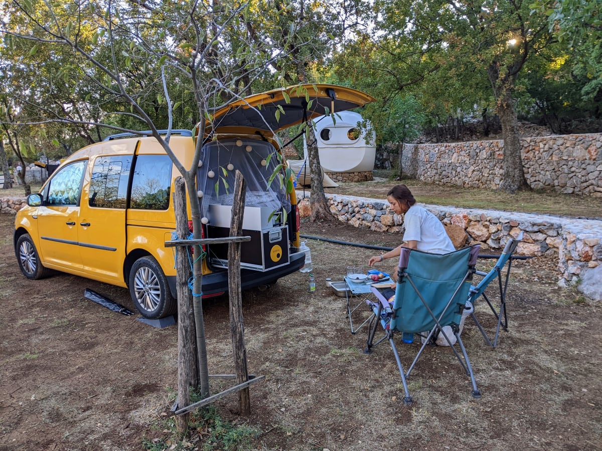 Heckküche offen, Bett aufgebaut, Susi beim Kochen unter dem Obstbaum.