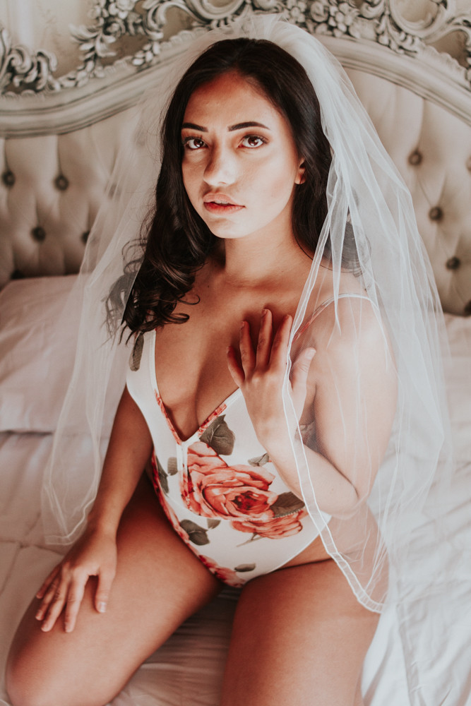 Model Gazing into the camera. She is wearing a wedding veil and a white bodysuit with floral print. 