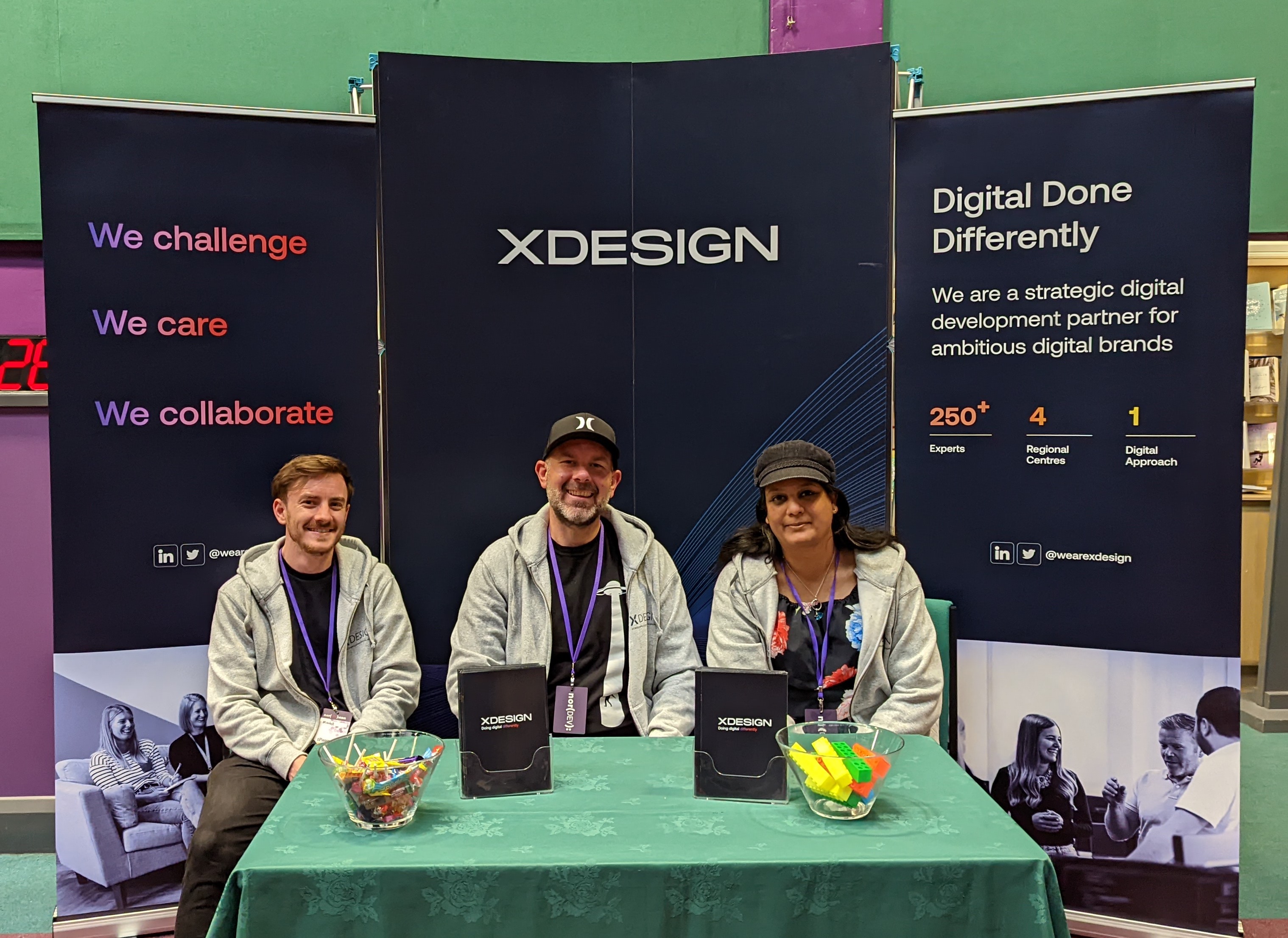 Three people sitting at an exhibition table at NorDevCon with xDesign banners behind them