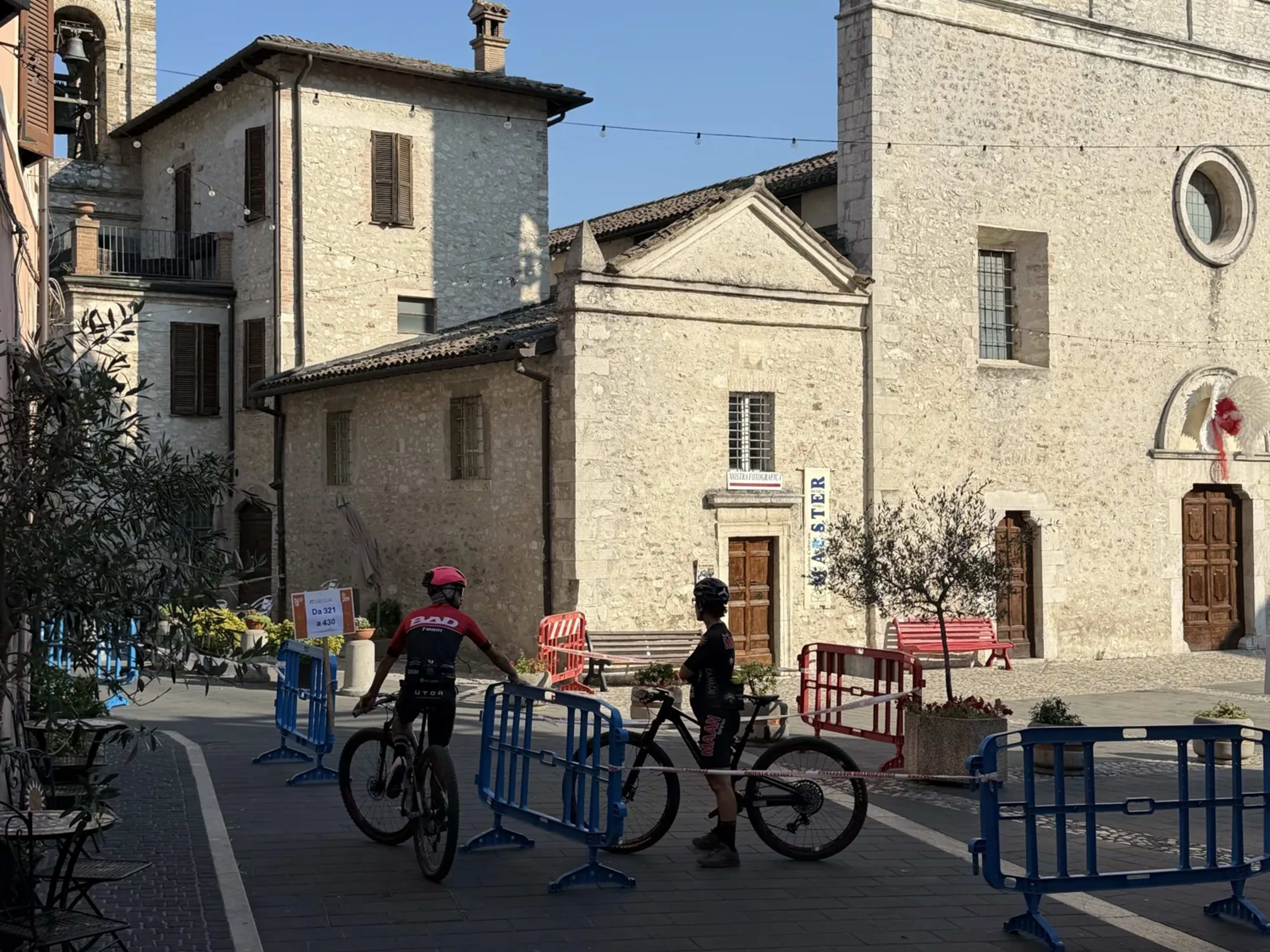 Due ciclisti in piazza davanti alla chiesa in pietra del borgo della Valnerina