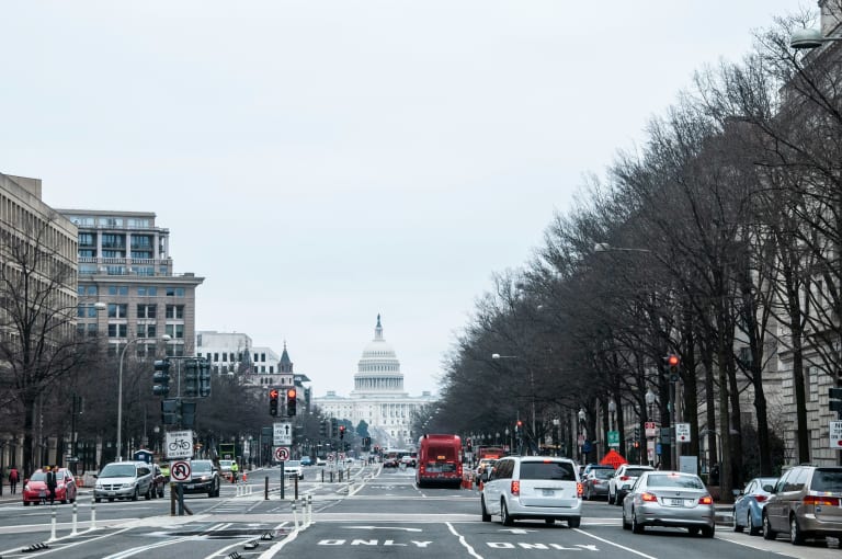 Vehicles driving on a city street during daytime with the U.S. Capitol building visible in the background.