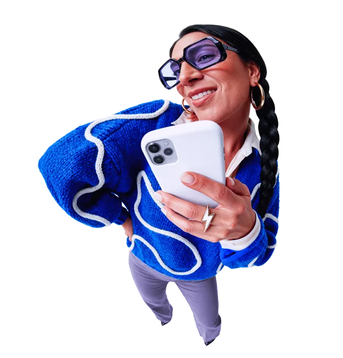 Woman holding a smartphone and smiling. She wears trendy sunglasses with purple collar lenses and a a c tricot blue sweater.
