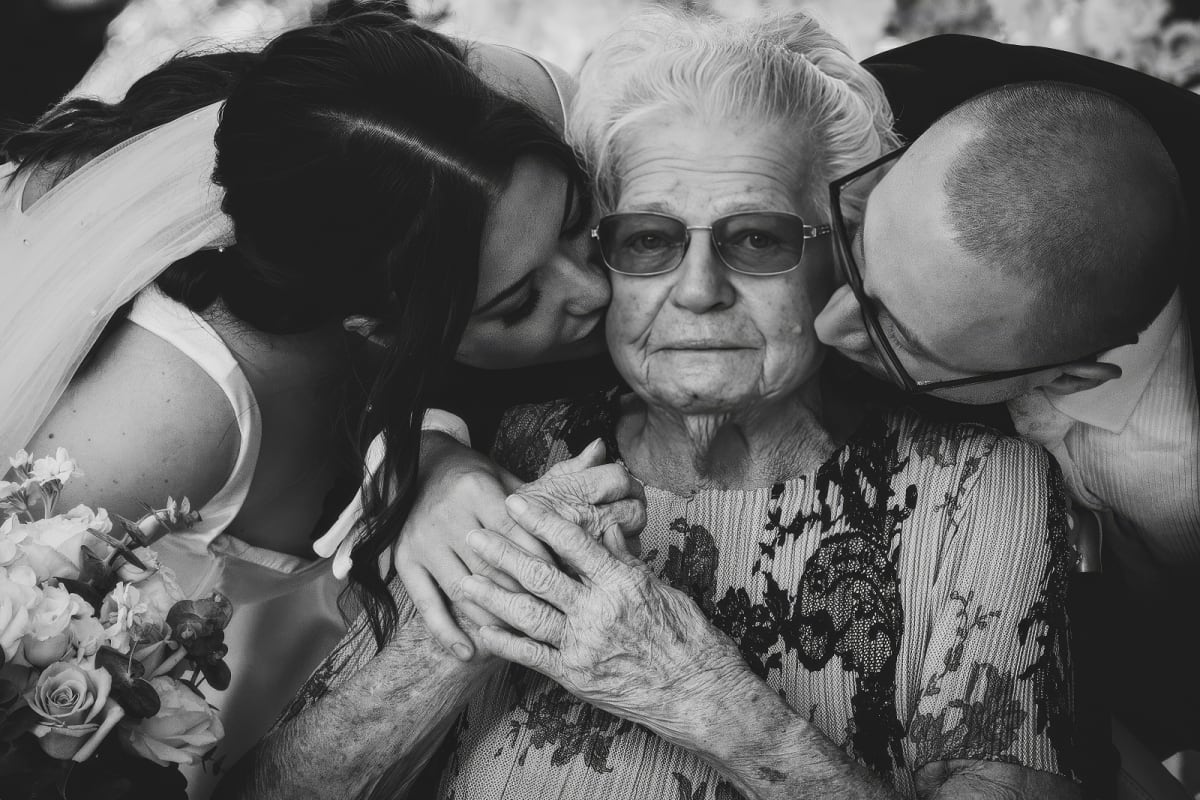 Black and white wedding photograph of bride and groom kissing grandmother on each cheek