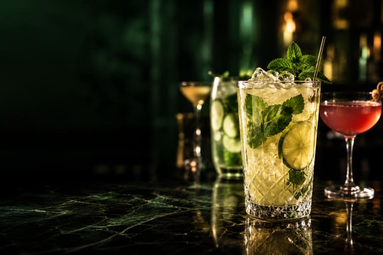 Drink alto com limão, hortelã e gelo picado sobre bancada de mármore verde, com outros coquetéis ao fundo