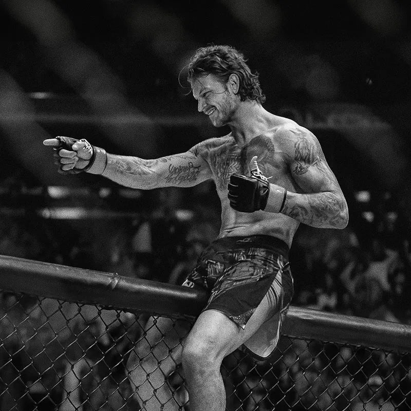 Tetovaný MMA bojovník s rukavicemi sedí na kleci a slaví vítězství před diváky.