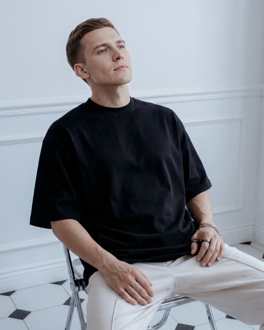 365.APParel Oversize T-shirt Black — man, seated on chair, looking up, three-quarter view, indoor editorial