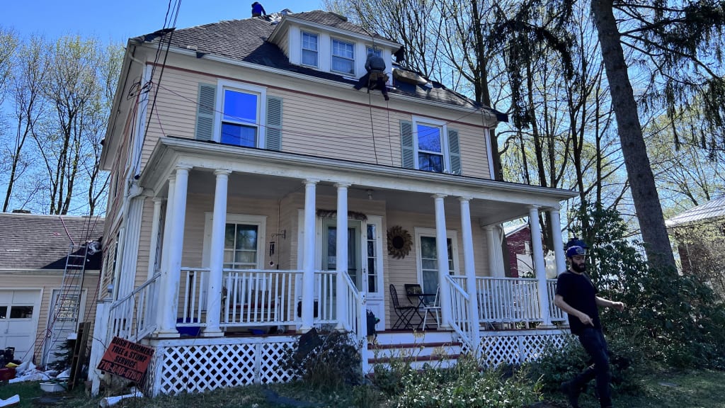 Two workers on a beige house roof with blue trim, one on the ground, clear sunny day.