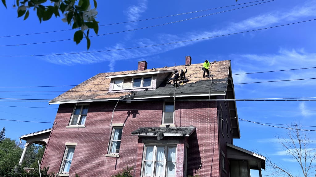 Roof repair workers in safety gear fixing shingles on a red brick house under clear sky.