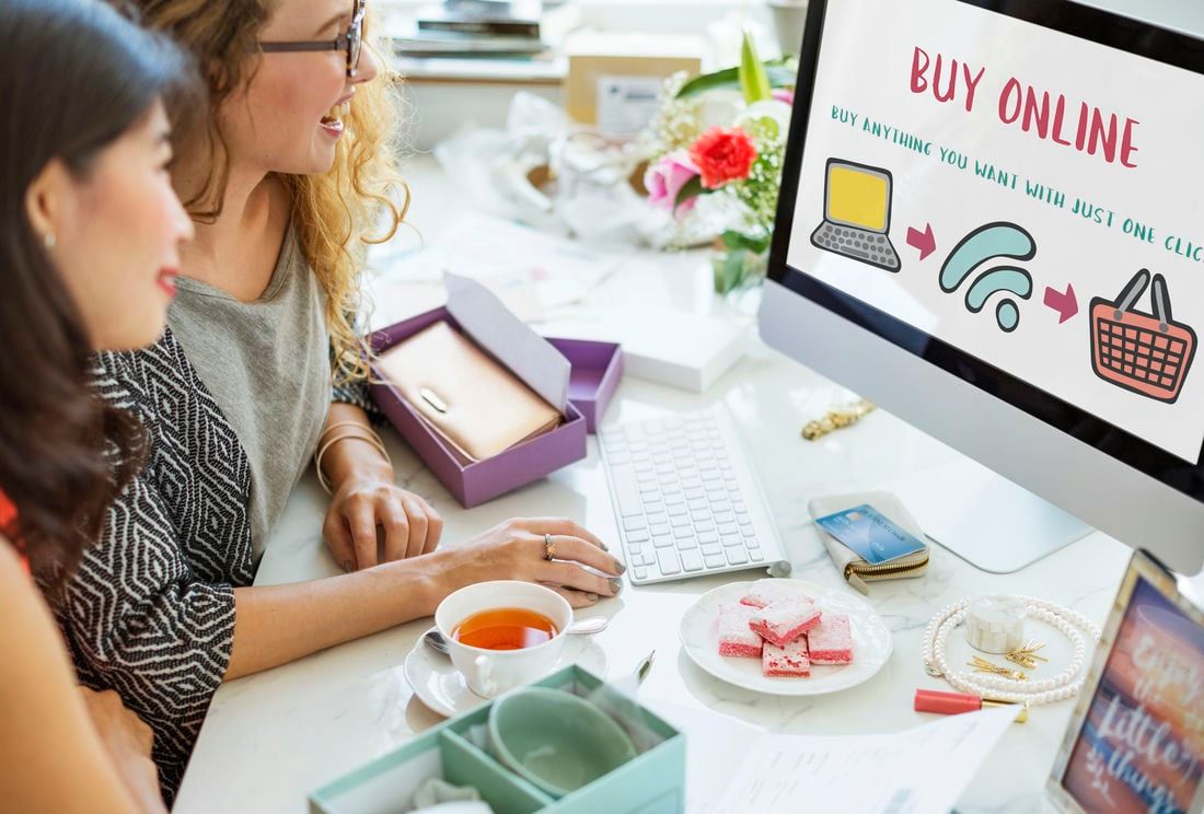 Two women enjoy online shopping together, exploring a website with a prominent "Buy Online" message, highlighting the convenience and appeal of e-commerce.