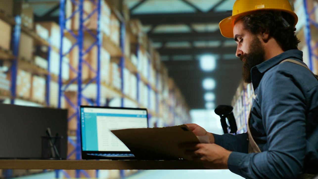 A warehouse worker reviews order details on a clipboard in front of a computer.