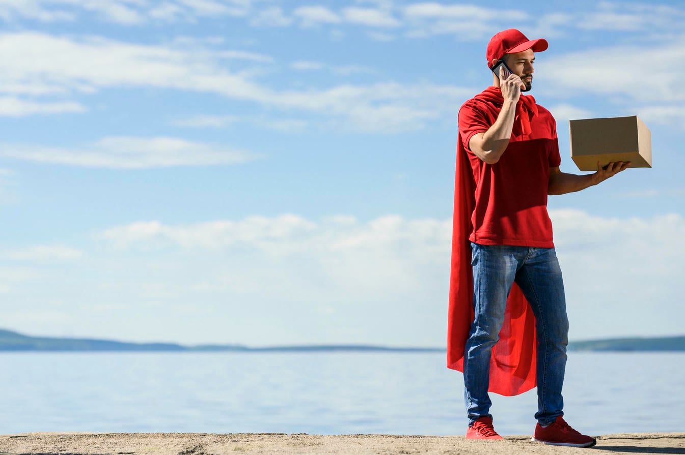 A delivery man in a superhero cape talks on the phone while holding a package by a lakeside.