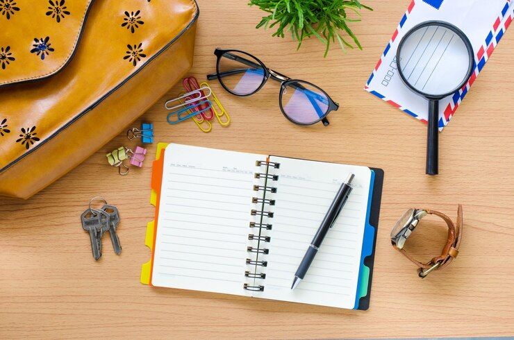 The image shows an open notebook surrounded by various items including a pen, keys, glasses, a magnifying glass, and other office supplies on a desk.