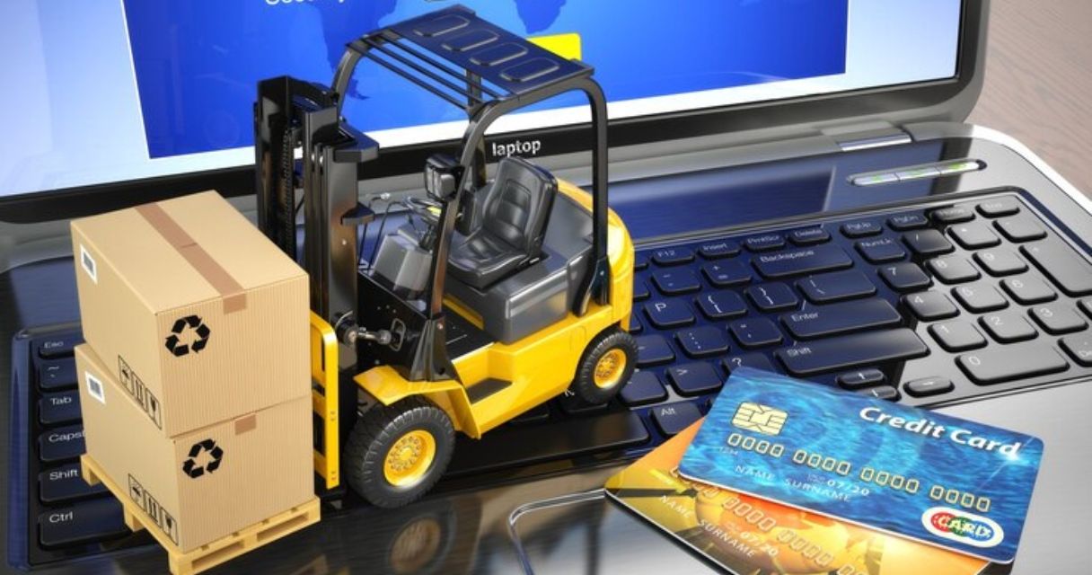 A miniature forklift carrying cardboard boxes on a laptop keyboard, with credit cards nearby, symbolizing e-commerce and online logistics.