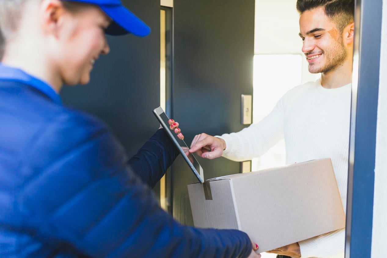 A delivery person handing a package to a recipient at the door, who is signing for the delivery on a tablet.