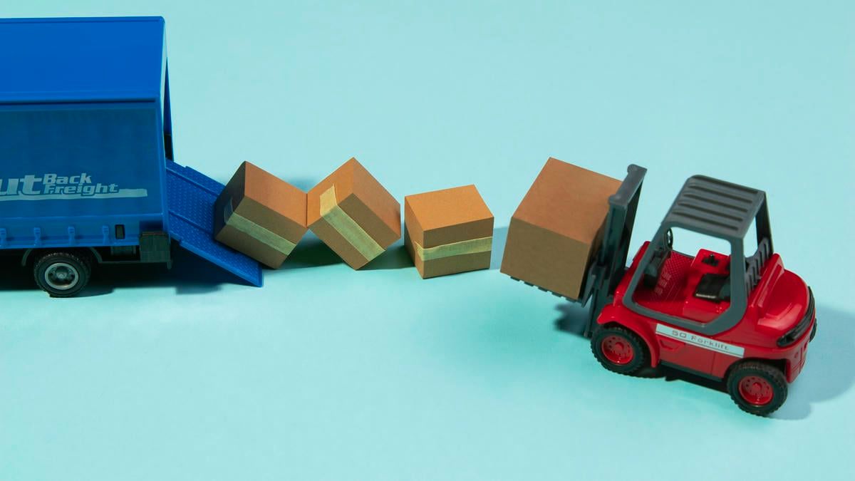 A toy forklift unloading cardboard boxes from a toy truck, illustrating the concept of logistics and transportation.