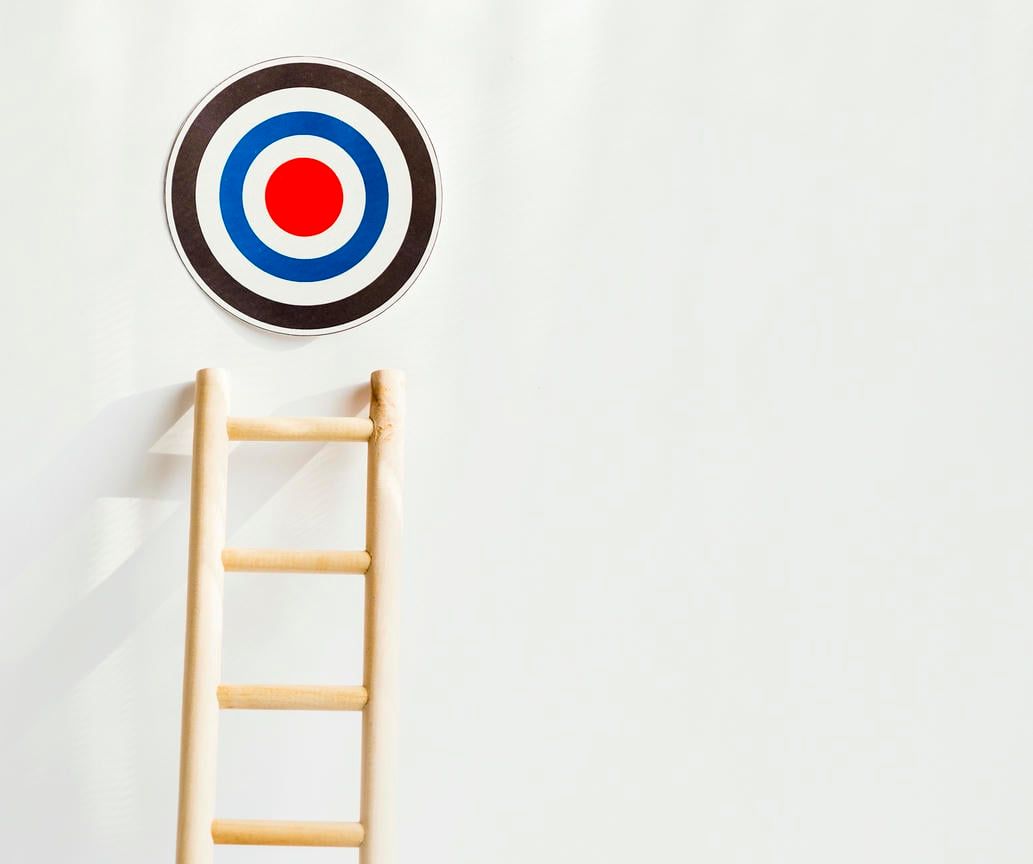 The image shows a wooden ladder positioned beneath a target on a white wall, symbolizing the steps or efforts needed to reach a goal.