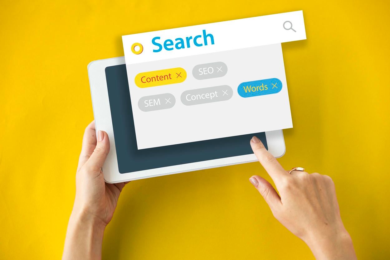 The image shows a person using a tablet to search for keywords related to content, SEO, SEM, and concepts, symbolizing the process of digital marketing and keyword research.