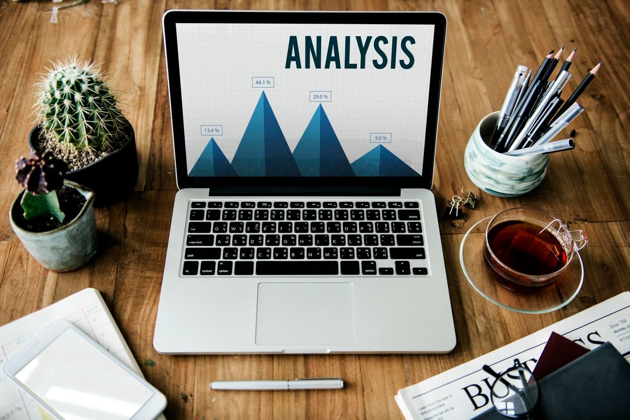 The image shows a laptop displaying an analysis graph, surrounded by cacti, pencils, a cup of tea, and business documents on a wooden desk, representing a workspace for data analysis and business planning.