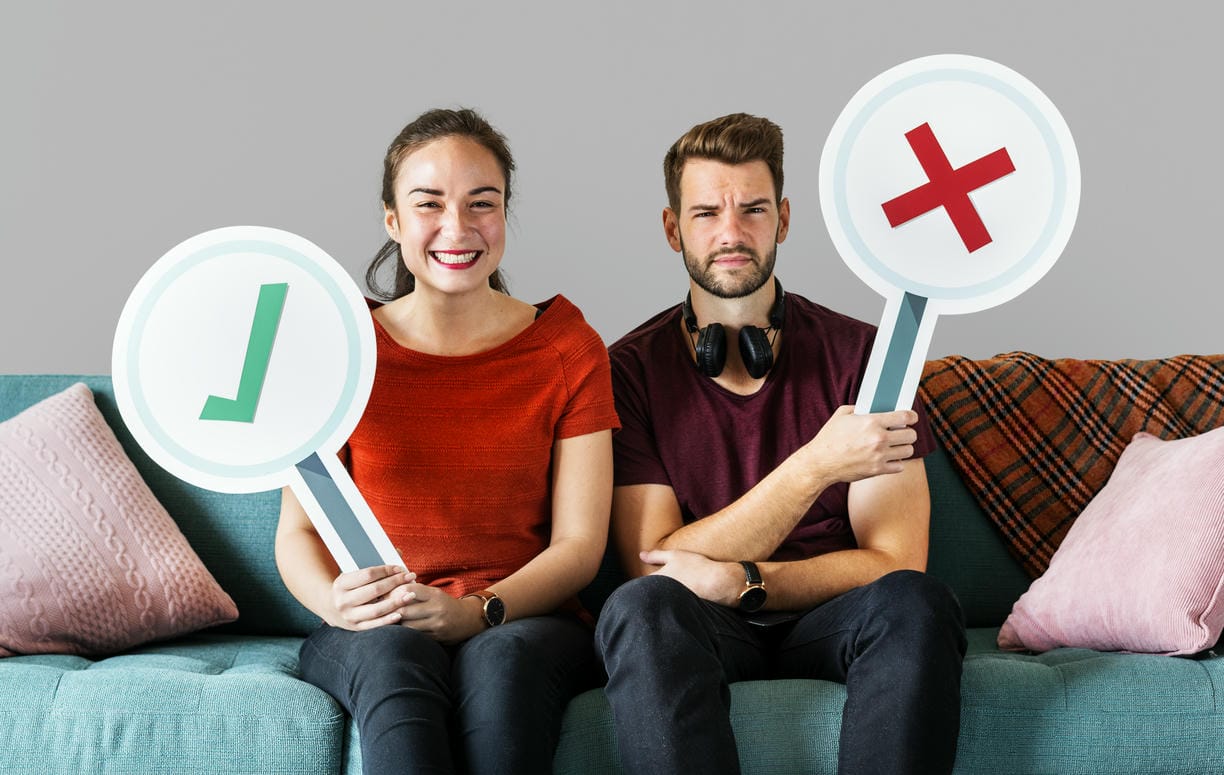 The image shows a couple on a couch holding signs with a check mark and a cross, respectively, likely expressing their differing opinions or preferences in a light-hearted manner.
