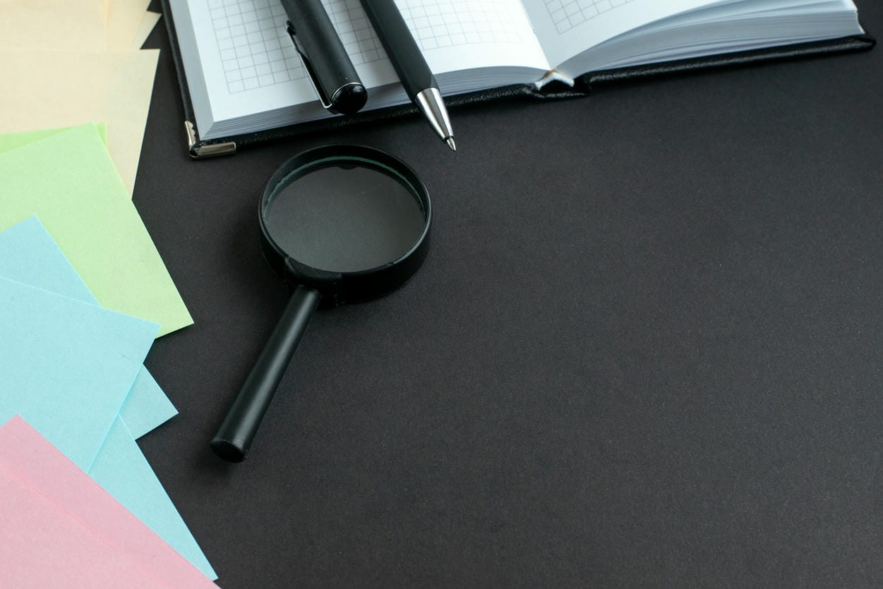 The image shows a workspace setup with a magnifying glass, colorful sticky notes, a pen, and an open notebook on a dark background, suggesting a scene of investigation or detailed study.