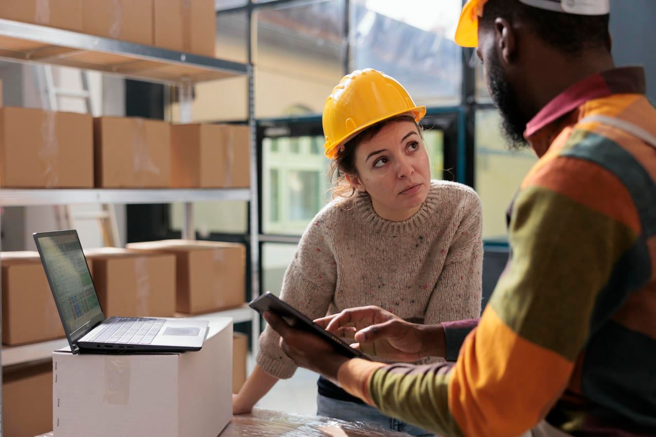 Two warehouse workers wearing safety helmets are discussing logistics while checking online orders on a tablet, with a laptop on a box and shelves of packages in the background.