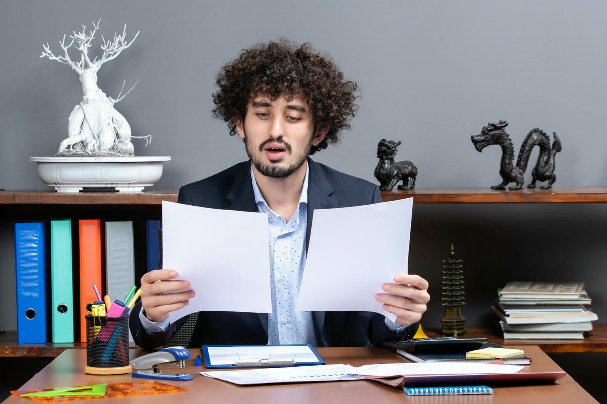 The image shows a young businessman with curly hair looking puzzled while holding and examining documents at his office desk, surrounded by various decorative objects and office supplies.