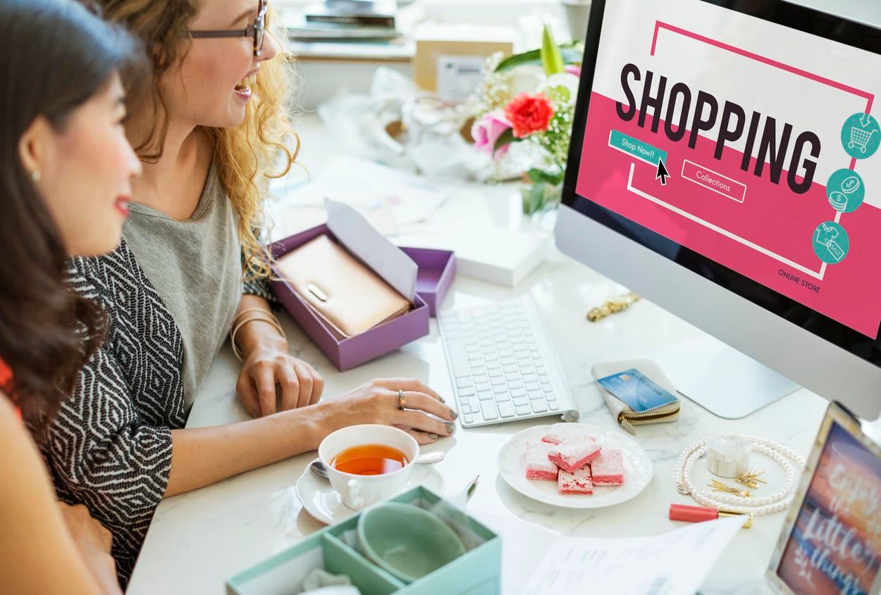 Two women are engaging in online shopping at a computer displaying a vibrant "SHOPPING" interface, surrounded by items like jewelry, desserts, and a cup of tea, creating a relaxed and consumer-focused atmosphere.