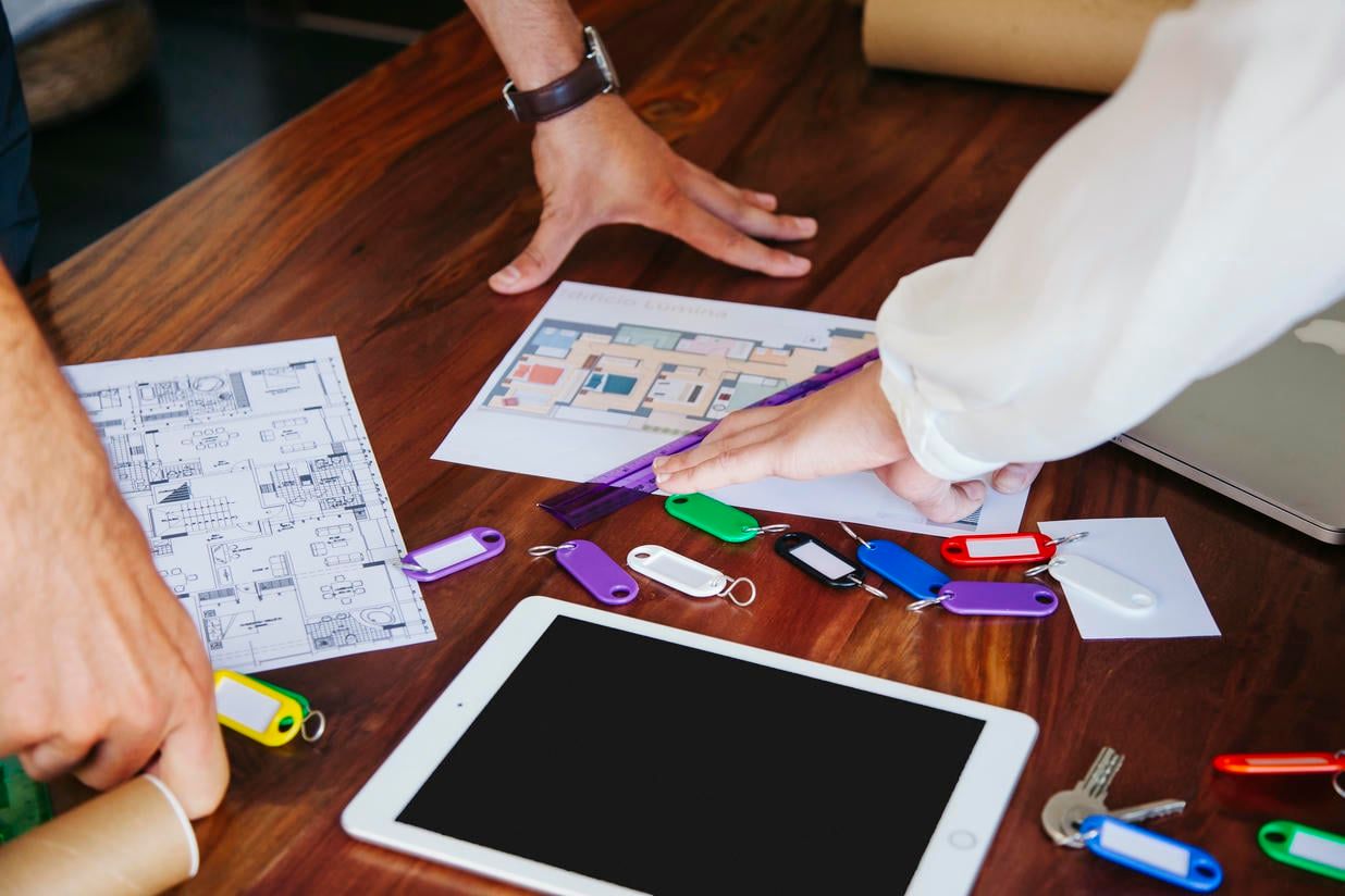 The image depicts two individuals reviewing architectural plans and key fobs on a wooden table, suggesting a real estate planning session.