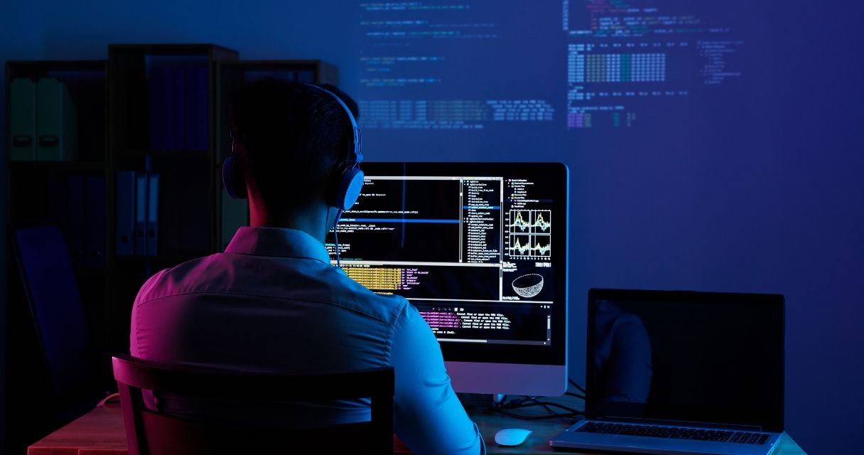 This image shows a programmer working late at night, intensely focused on coding tasks displayed across multiple computer screens in a dimly lit room.