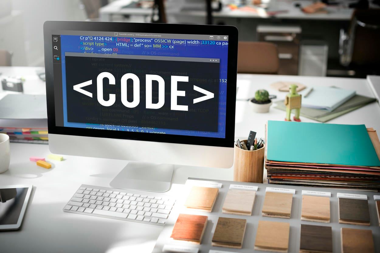 The image shows a computer screen with "CODE" displayed, surrounded by design materials in a creative workspace.
