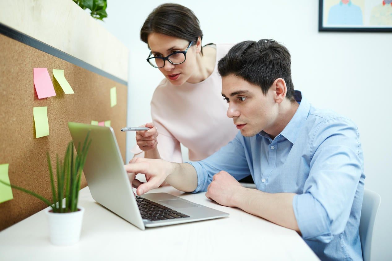 The image shows two people collaborating at a desk, with one person pointing at a laptop screen while the other looks on attentively, surrounded by sticky notes on a corkboard, suggesting a brainstorming or planning session.
