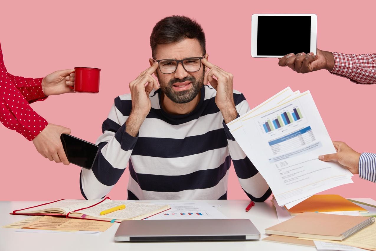 The image shows a man sitting at a desk, looking stressed and overwhelmed, as multiple hands present him with various items like gadgets, papers, and a coffee cup, symbolizing multitasking or information overload.