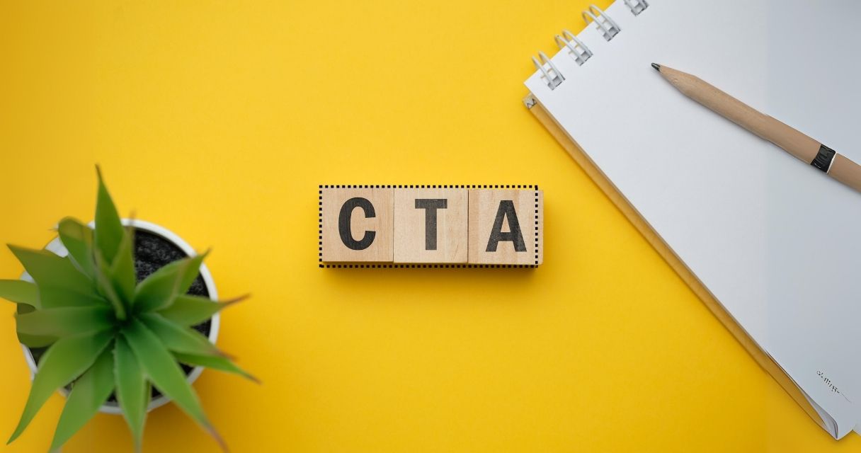 The image shows a creative flat lay with a wooden block spelling out "CTA," flanked by a green plant and a pencil on a notebook, all set against a bright yellow background, symbolizing the concept of "Call to Action" in marketing and design.
