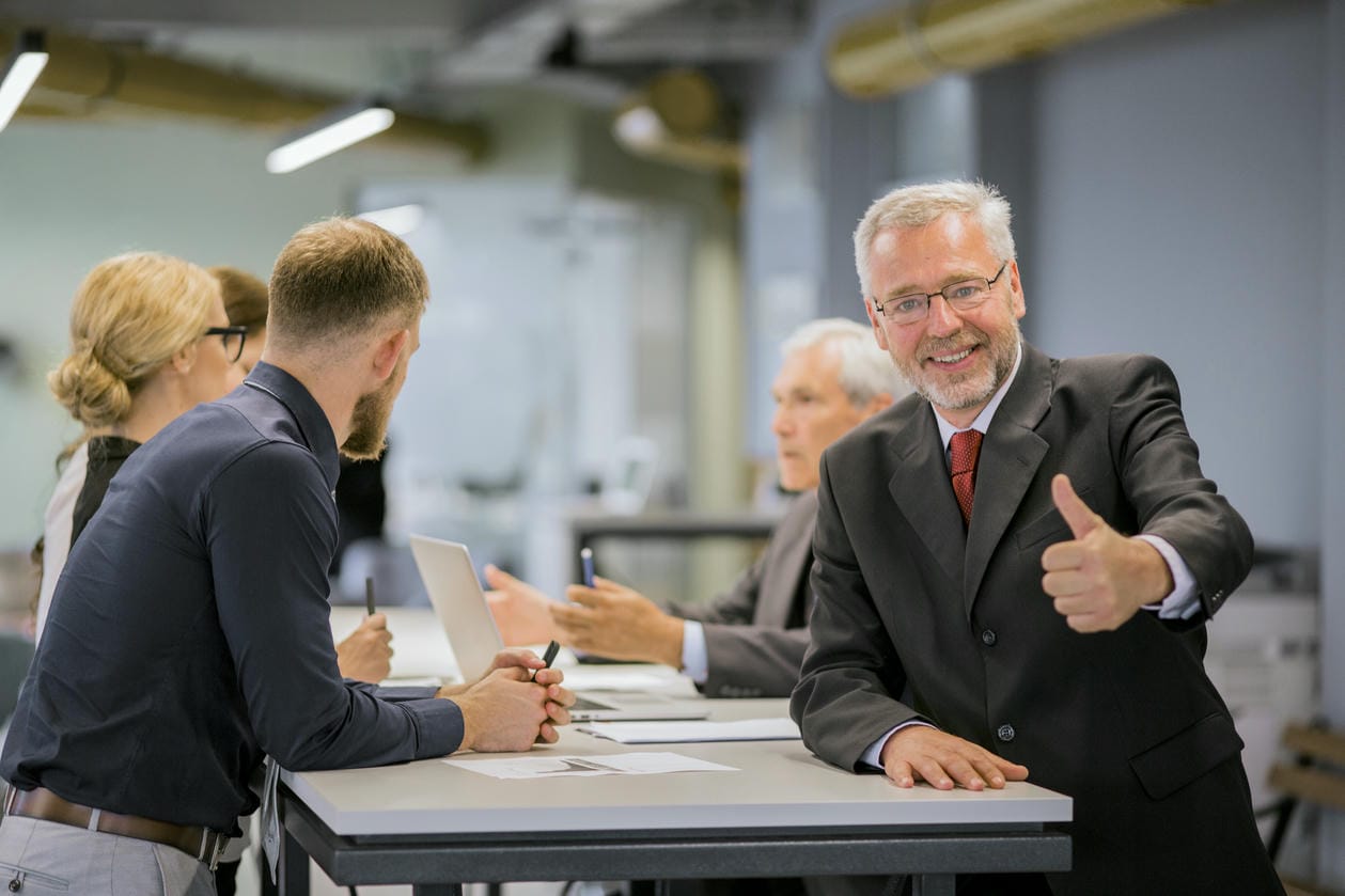 Smiling senior businessman showing thumb up sign in front of businesspeople discussing in the office