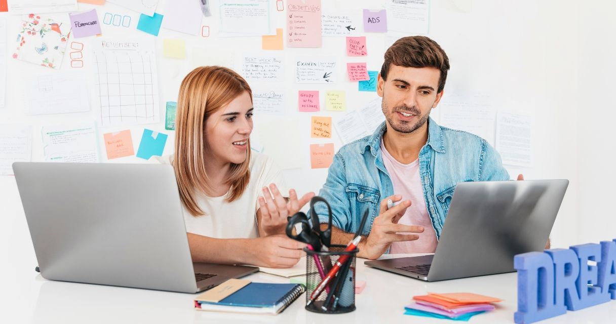 Smiling male and female sitting at desk and talking looking at laptop