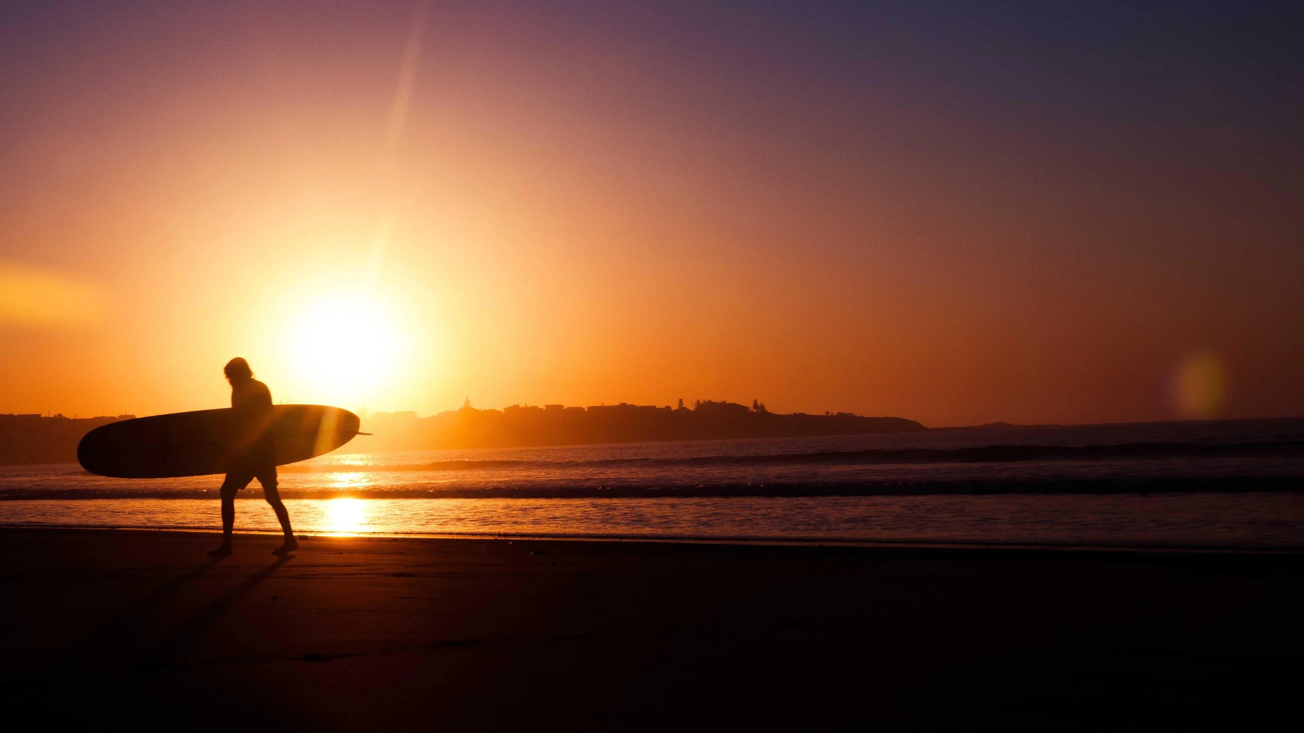 A surfer carrying a board along the beach at sunset
