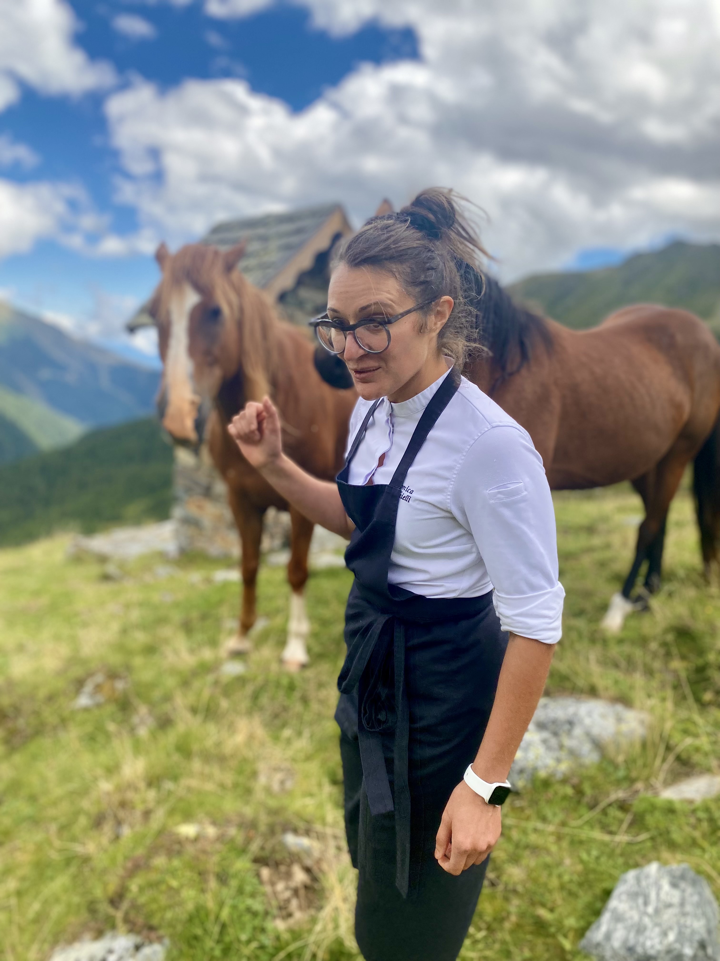 Veronica Forchielli in tenuta da chef tra i pascoli in quota, con due cavalli al pascolo sullo sfondo
