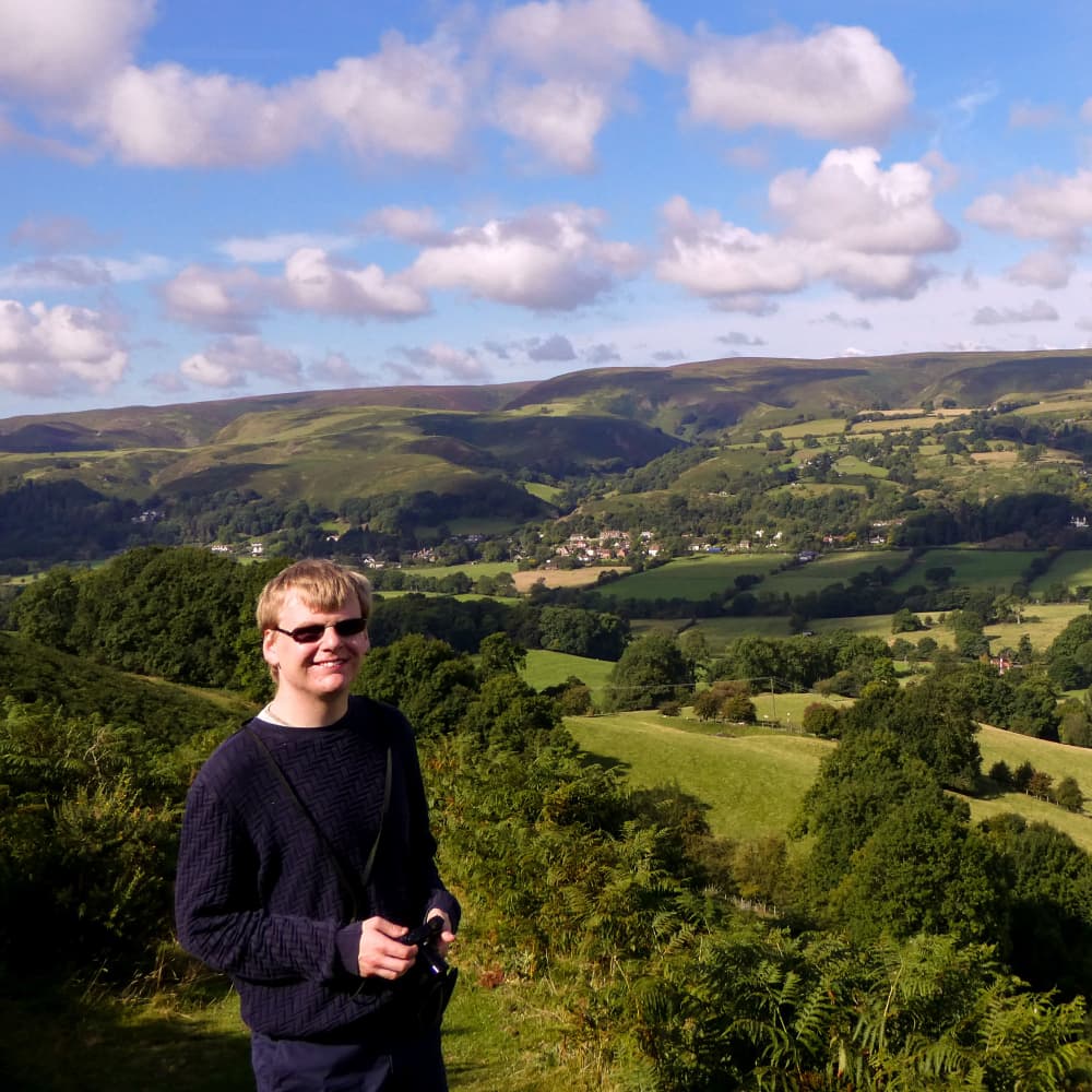 Duncan Ritchie smiling while holding his camera, surrounded by sunlit hills