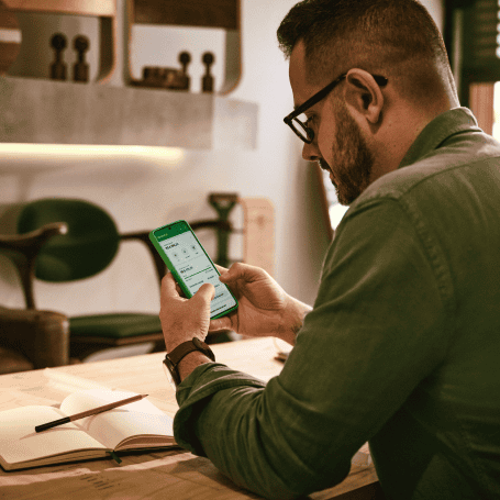 Empreendedor sentado e apoiado com os braços na mesa em sua loja, segurando o celular com o aplicativo da Stone aberto. Na mesa um caderno com lápis. E ao fundo cadeira e decorações.