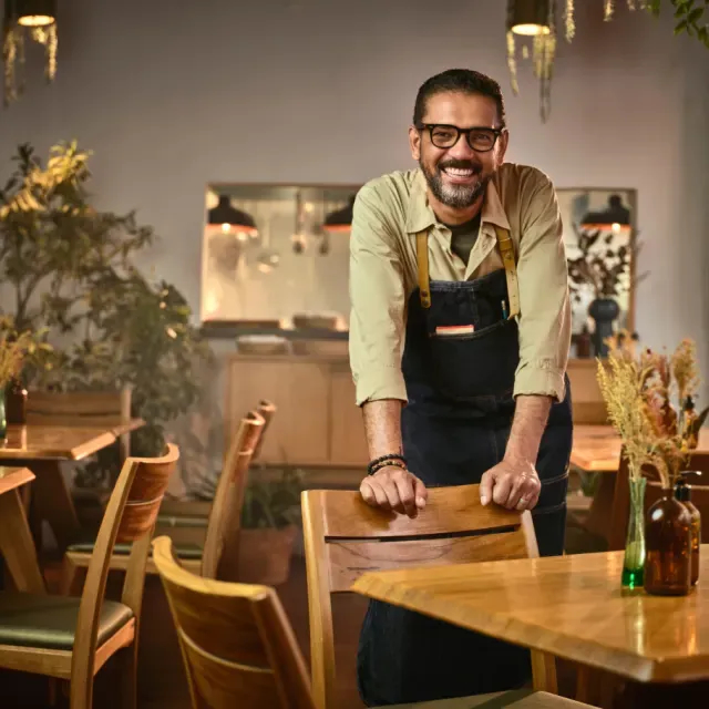 Empreendedor com avental no seu restaurante sorrindo para a foto apoiado na cadeira.