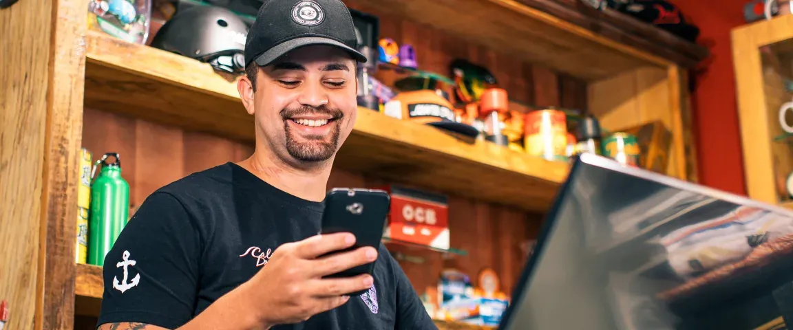 Empreendedor em sua loja olhando para o celular e ao canto a sua Maquina da Stone