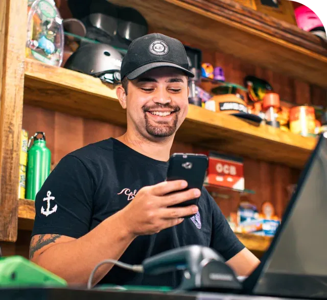 Empreendedor em sua loja olhando para o celular e ao canto a sua Maquina da Stone