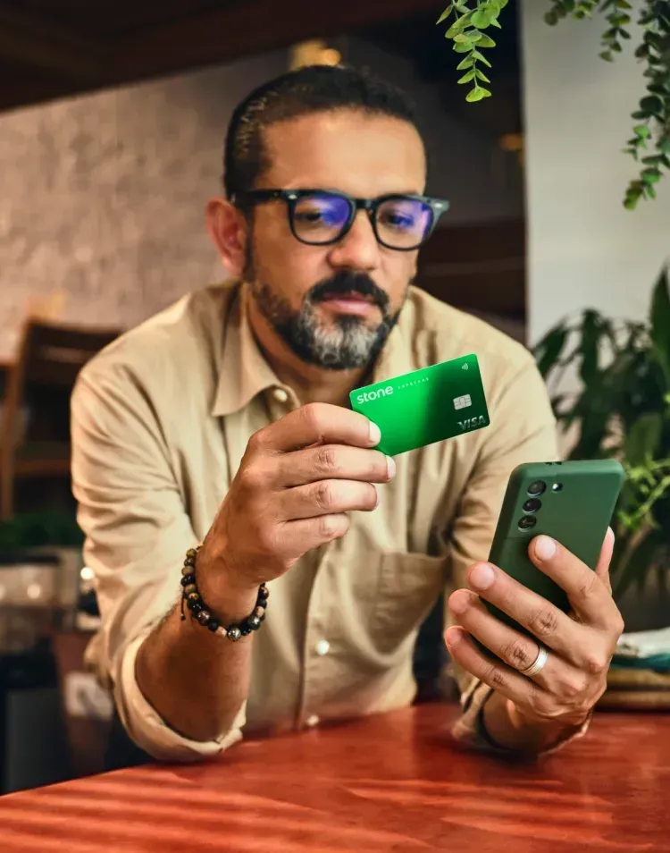 Empreendedor apoiado no balcão segurando o seu celular em uma mão e na outra o seu cartão Stone