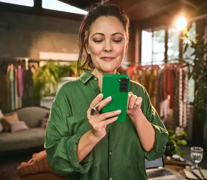 Empreendedora em sua loja segurando o celular. Ao fundo as roupas penduradas em araras e plantas como decorações.