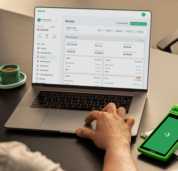 Empreendedor utilizando o notebook em cima da mesa com a máquina de cartão Stone ao lado