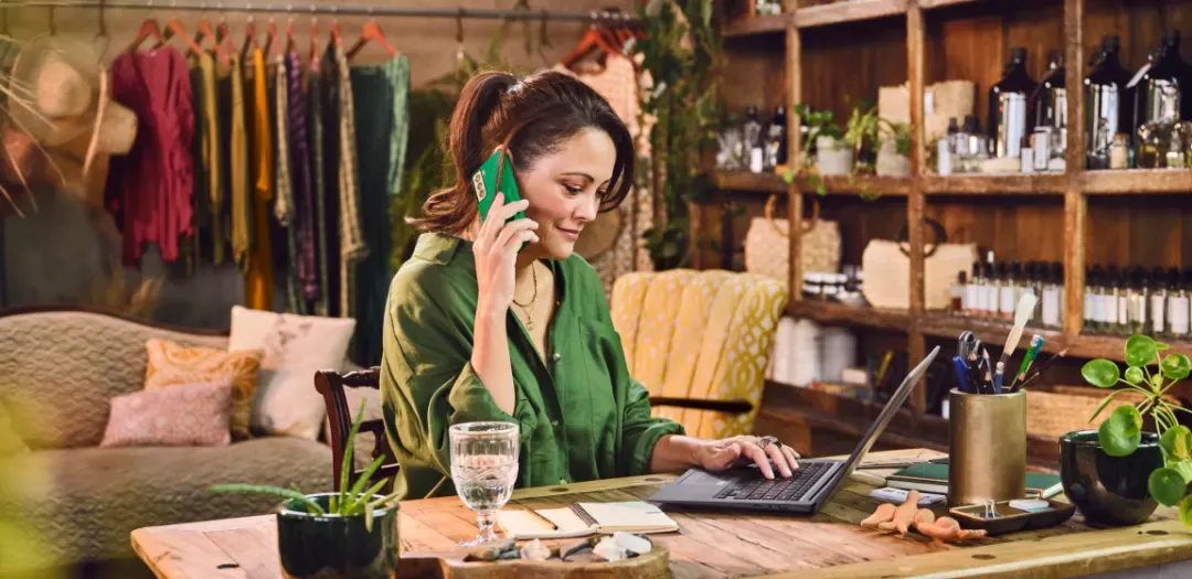 Empreendedora em sua loja utilizando o notebook e falando ao telefone. No balcão consta um copo de água, vaso de planta e porta lápis. E ao fundo peças de roupas penduradas na arara e nas prateleiras bolsas e frascos.