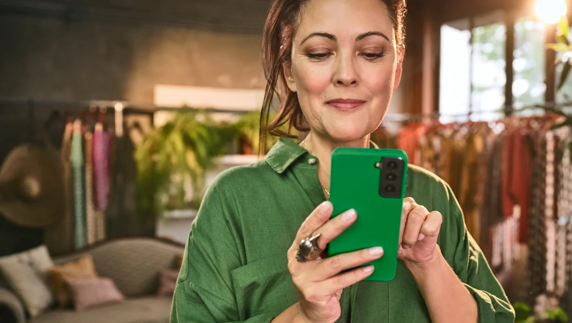 Empreendedora em sua loja segurando o celular. Ao fundo as roupas penduradas em araras e plantas como decorações.