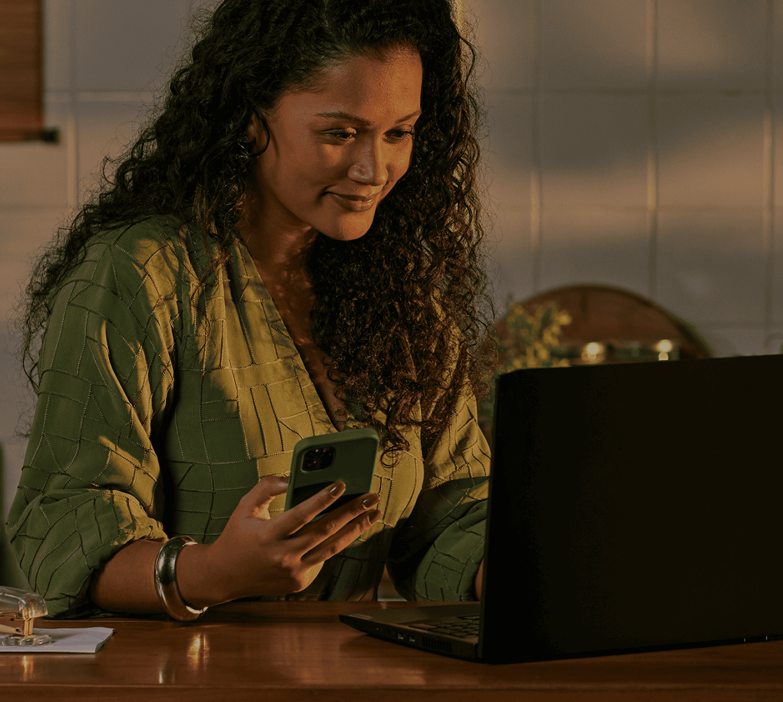 Empreendedora apoiada no balcão da sua cozinha, utilizando o seu notebook e segurando o celular. 