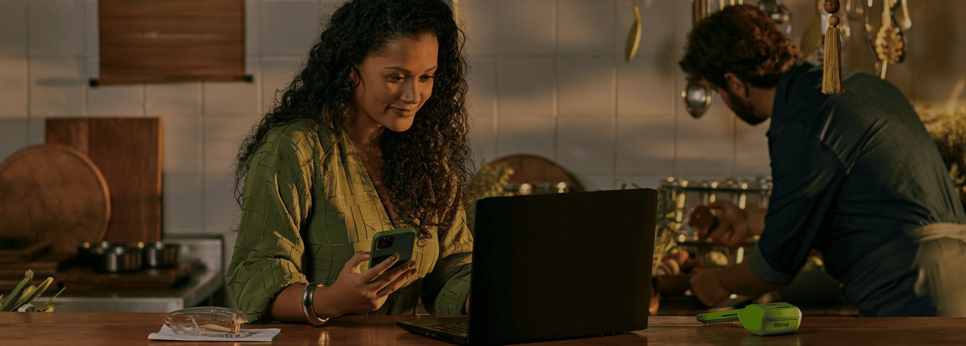 Empreendedora apoiada no balcão da sua cozinha, utilizando o seu notebook e segurando o celular. A Maquininha de Cartão Stone e um grampeador também está em cima do balcão. Ao fundo o cozinheiro está segurando uma panela e tempero. 