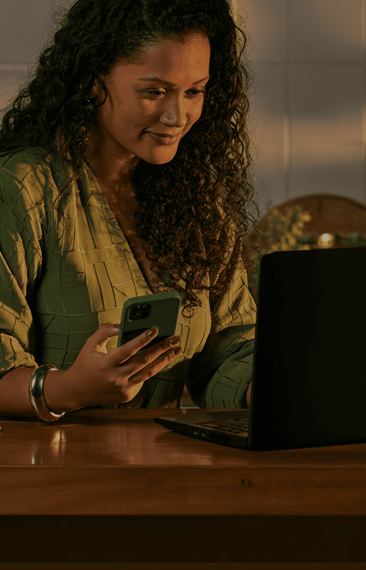 Empreendedora de um estabelecimento culinário com celular na mão, apoiada na mesa com seu notebook e uma maquininha de cartão da Stone ao seu lado. E ao fundo, um cozinheiro na frente do balcão com utensílios de cozinha.